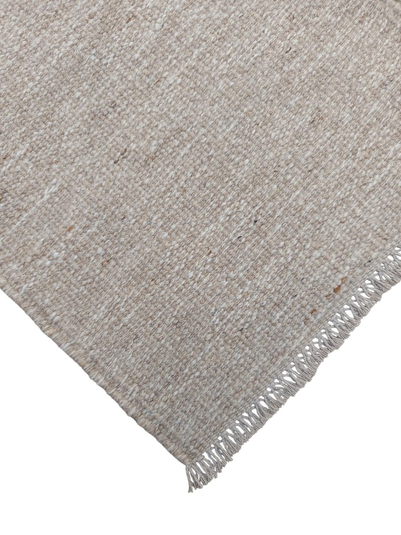 K&ouml;nnte beinhalten: Ein rechteckiger Webteppich in einer strukturierten, neutralen Farbpalette aus Beige und Off-White. Der Stoff zeigt ein dichtes, geschlungenes Webmuster mit dezenten braunen Sprenkeln. Eine Kante ist mit einer kurzen, geraden Franse versehen.