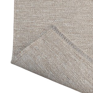 K&ouml;nnte beinhalten: Ein rechteckiger Teppich mit gewebter Textur in einer neutralen Farbpalette aus Beige und Off-White. Der Teppich hat eine umgeschlagene Ecke, die die Unterseite freigibt und eine sichtbare Fransenkante an der kurzen Seite des Gewebes zeigt.