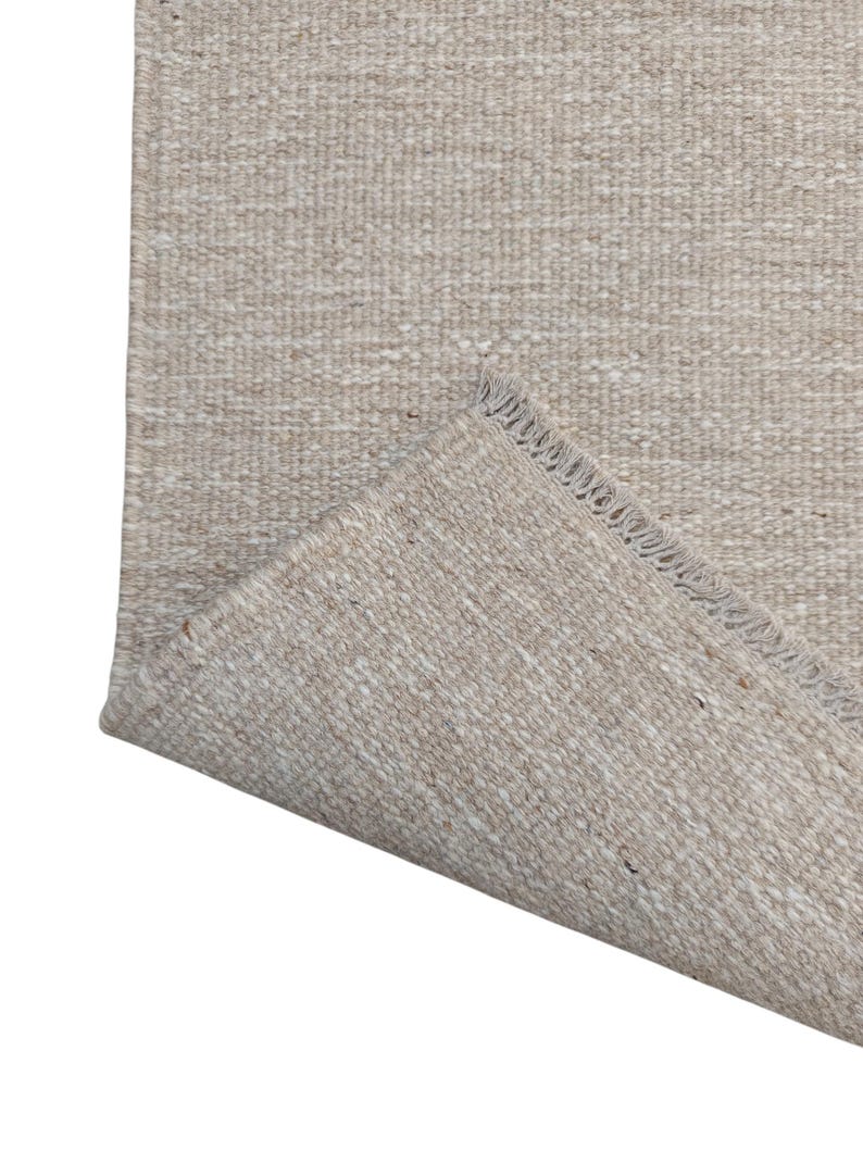 K&ouml;nnte beinhalten: Ein rechteckiger Teppich mit gewebter Textur in einer neutralen Farbpalette aus Beige und Off-White. Der Teppich hat eine umgeschlagene Ecke, die die Unterseite freigibt und eine sichtbare Fransenkante an der kurzen Seite des Gewebes zeigt.