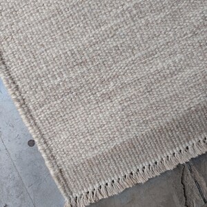 Pu&ograve; includere: Un tappeto rettangolare caratterizzato da una trama tessuta in una palette di colori neutri beige e crema. Il tappeto presenta una sezione di bordo distinta con un motivo a trama pi&ugrave; stretta e una frangia composta da corte nappe annodate.