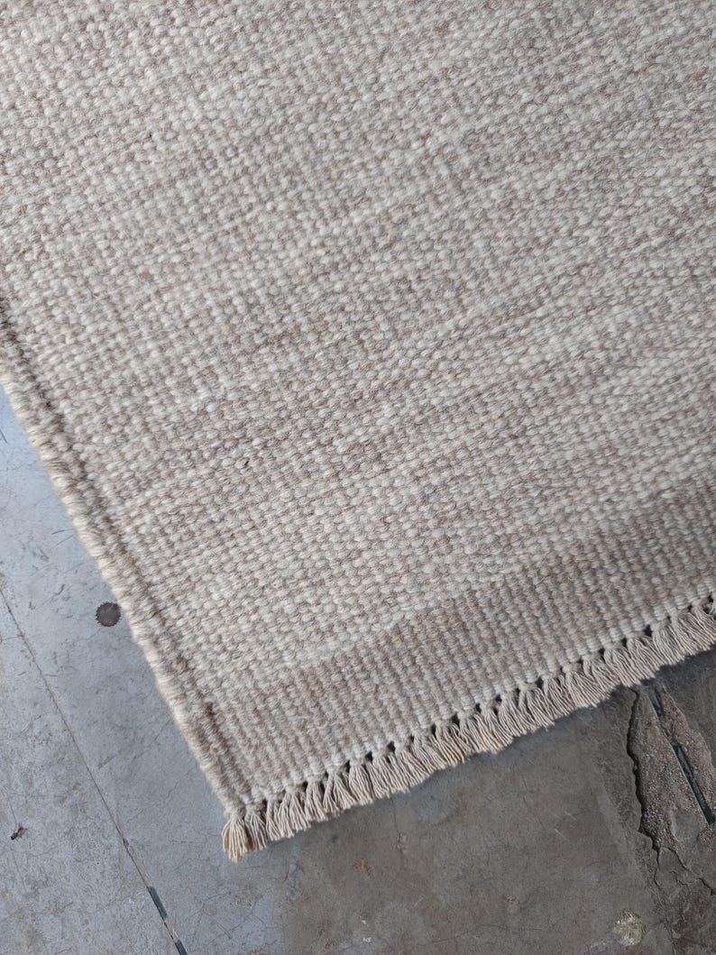 Pu&ograve; includere: Un tappeto rettangolare caratterizzato da una trama tessuta in una palette di colori neutri beige e crema. Il tappeto presenta una sezione di bordo distinta con un motivo a trama pi&ugrave; stretta e una frangia composta da corte nappe annodate.