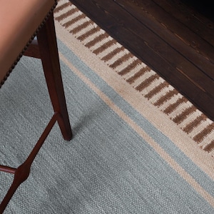 Puede incluir: Una alfombra tejida que presenta un gran campo central azul claro bordeado por una fina franja beige y una banda más ancha de rayas verticales alternas de color marrón oscuro y crema, parcialmente cubierta por la pata de madera de una silla de cuero beige.