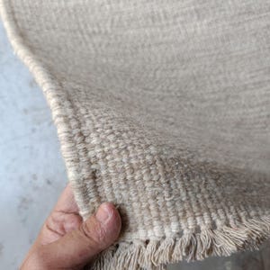 Pu&ograve; includere: Un tappeto intrecciato color beige con una superficie testurizzata e un bordo sfrangiato. Il tappeto presenta una costruzione densa e ad anello realizzata in fibre naturali, mostrando un colore neutro costante e un bordo dritto e rifinito.