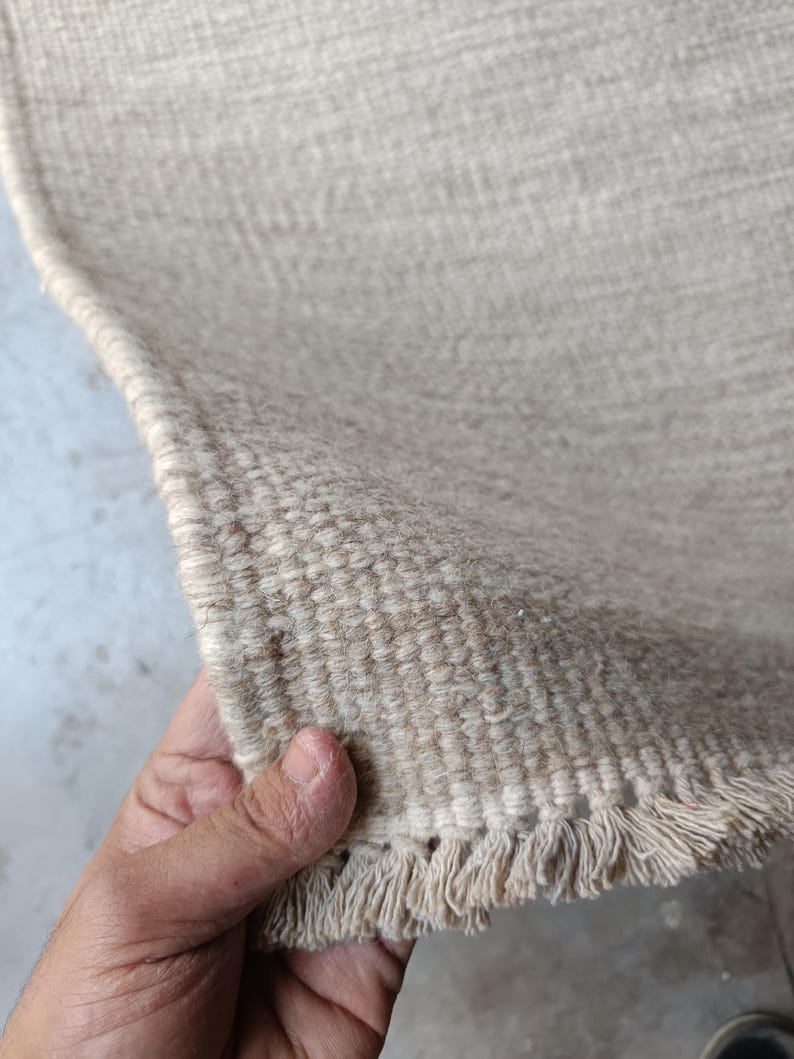 Pu&ograve; includere: Un tappeto intrecciato color beige con una superficie testurizzata e un bordo sfrangiato. Il tappeto presenta una costruzione densa e ad anello realizzata in fibre naturali, mostrando un colore neutro costante e un bordo dritto e rifinito.