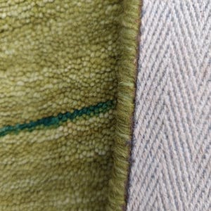 May include: A close-up view of a textured lime green rug with a dark green horizontal stripe. The rug features a thick, looped pile construction and a finished edge with a visible light gray herringbone pattern on the underside backing material.