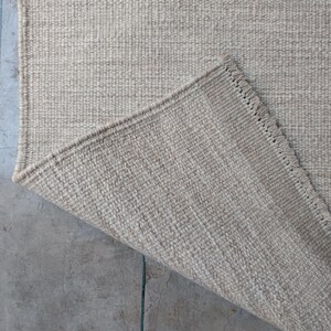 Pu&ograve; includere: Un tappeto intrecciato in tonalit&agrave; beige neutra caratterizzato da una costruzione a pelo riccio denso e testurizzato. Un angolo del tappeto &egrave; ripiegato per rivelare il retro, mostrando un bordo rifinito con frange corte e uniformi.