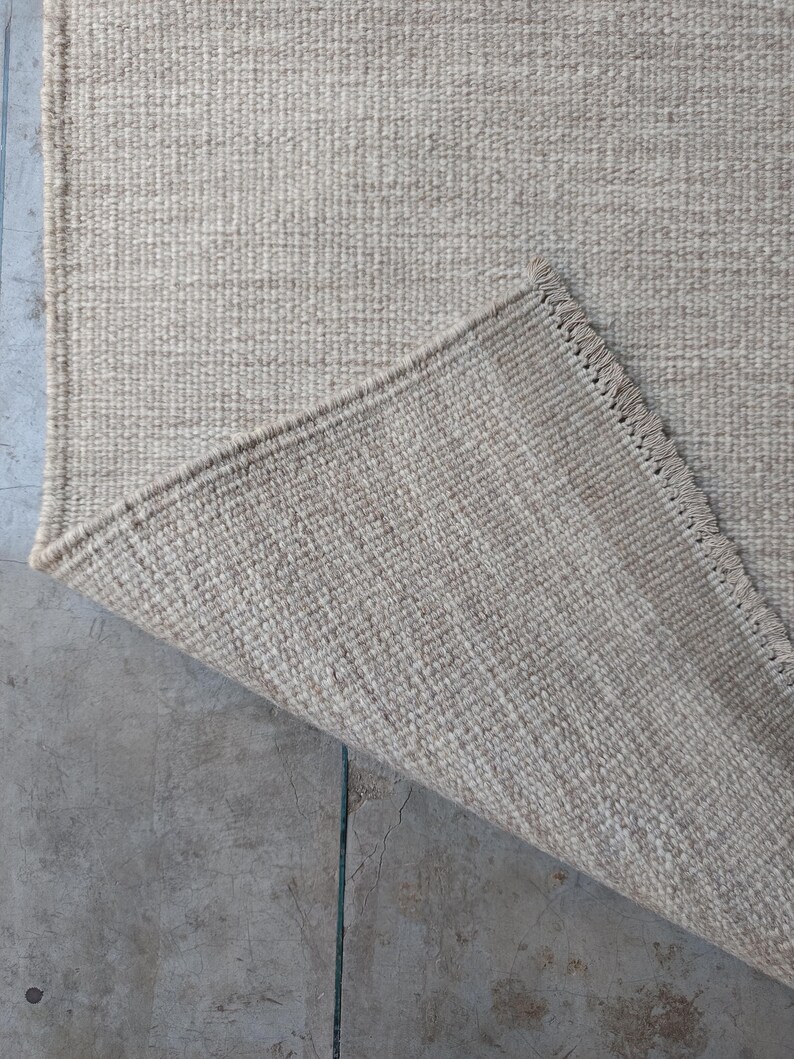 Pu&ograve; includere: Un tappeto intrecciato in tonalit&agrave; beige neutra caratterizzato da una costruzione a pelo riccio denso e testurizzato. Un angolo del tappeto &egrave; ripiegato per rivelare il retro, mostrando un bordo rifinito con frange corte e uniformi.