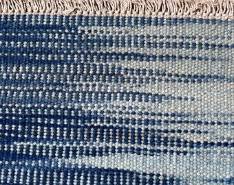 May include: Close-up of a blue and white patterned rug. The rug has a textured surface with a fringe along the top edge. The design features vertical stripes and a mix of light and dark blue hues.