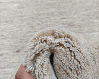 May include: A thick, beige, high-pile rug with a soft, shaggy texture. The material is folded over to reveal the dense, plush fibers and the construction of the pile. The surface has a uniform, neutral light tan color throughout the entire piece.