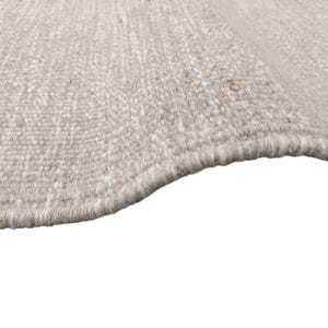 K&ouml;nnte beinhalten: Ein gewebter Teppich mit einer strukturierten Oberfl&auml;che aus einer Mischung von beigen, hellbraunen und cremewei&szlig;en Fasern. Der Teppich hat eine dicke Schlingenflor-Konstruktion mit einer leicht geschwungenen Kante und dichtem Garn.