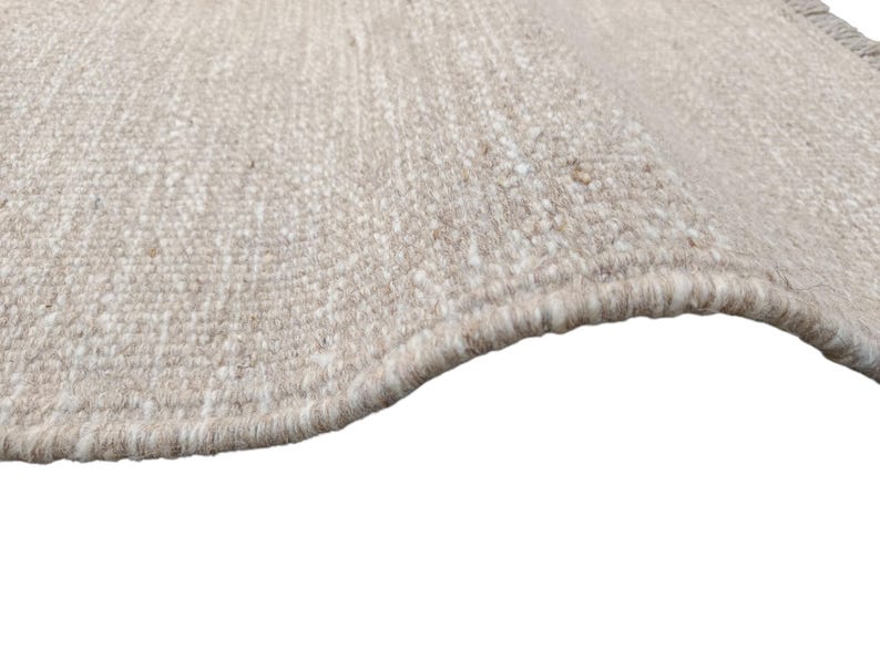 K&ouml;nnte beinhalten: Ein gewebter Teppich mit einer strukturierten Oberfl&auml;che aus einer Mischung von beigen, hellbraunen und cremewei&szlig;en Fasern. Der Teppich hat eine dicke Schlingenflor-Konstruktion mit einer leicht geschwungenen Kante und dichtem Garn.