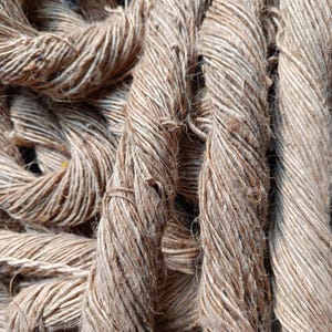 May include: A collection of thick, light brown twisted jute ropes with a coarse, fibrous texture. The strands are tightly wound together, creating a rugged, multi-layered appearance with visible loose fibers and a natural, earthy tan color throughout.