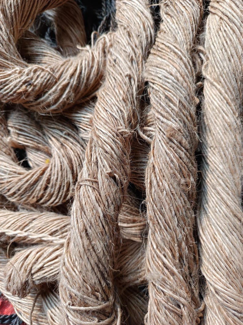 May include: A collection of thick, light brown twisted jute ropes with a coarse, fibrous texture. The strands are tightly wound together, creating a rugged, multi-layered appearance with visible loose fibers and a natural, earthy tan color throughout.