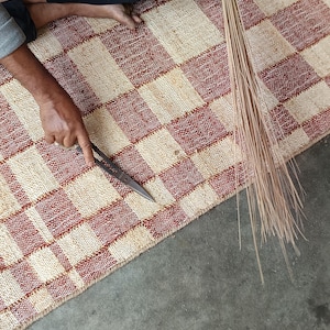 May include: A woven rug featuring a checkered pattern in alternating squares of natural beige and deep red. The rug is being trimmed with a pair of metal scissors, and a bundle of thin, light brown natural fibers is held alongside the edge of the rug.