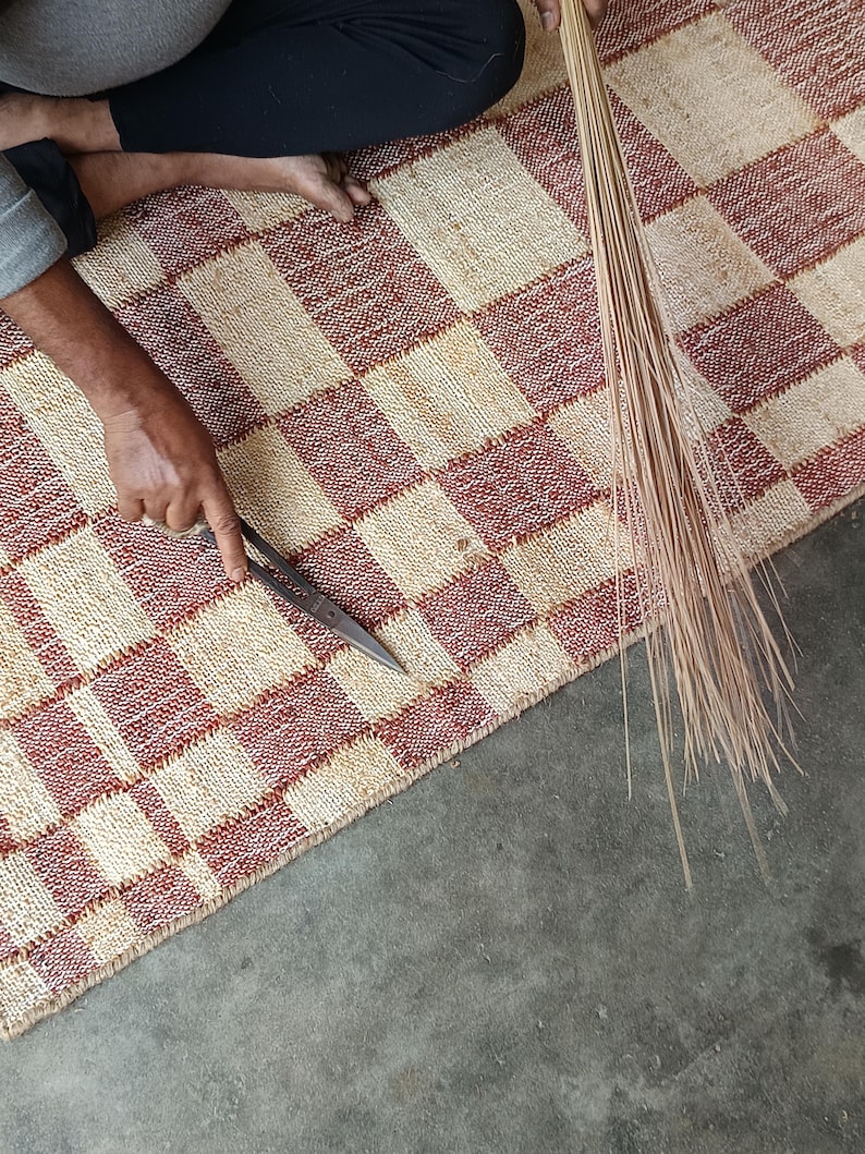 May include: A woven rug featuring a checkered pattern in alternating squares of natural beige and deep red. The rug is being trimmed with a pair of metal scissors, and a bundle of thin, light brown natural fibers is held alongside the edge of the rug.