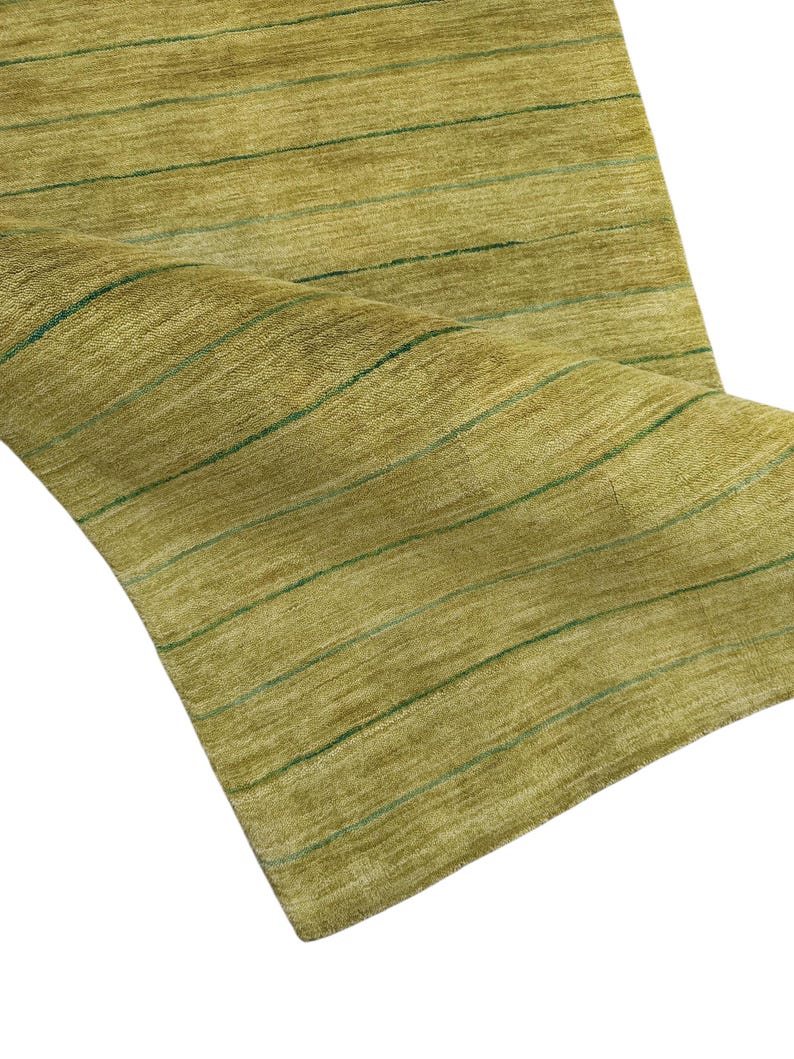 May include: A rectangular area rug featuring a textured olive green surface with thin, horizontal dark green stripes running across the length. The rug is partially folded over, revealing the soft, dense pile and the consistent linear pattern.
