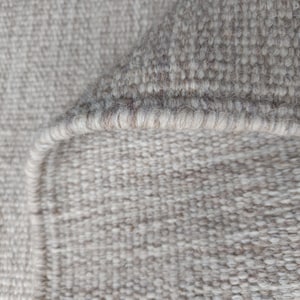 Pu&ograve; includere: Una vista ravvicinata di un tappeto intrecciato caratterizzato da una palette di colori beige neutro e bianco sporco. La trama mostra una densa costruzione a ricciolo con un bordo rifinito visibile dove il tappeto &egrave; ripiegato su se stesso.