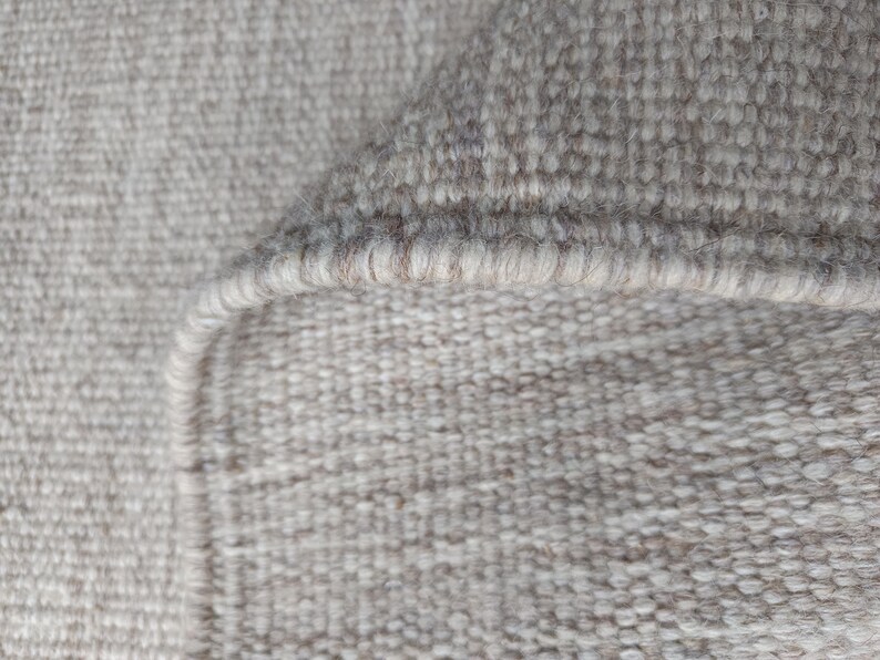 Pu&ograve; includere: Una vista ravvicinata di un tappeto intrecciato caratterizzato da una palette di colori beige neutro e bianco sporco. La trama mostra una densa costruzione a ricciolo con un bordo rifinito visibile dove il tappeto &egrave; ripiegato su se stesso.