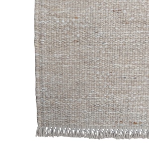 K&ouml;nnte beinhalten: Ein rechteckiger Webteppich in einer neutralen Farbpalette aus Beige und Off-White. Die Oberfl&auml;che zeigt ein gleichm&auml;&szlig;iges, dichtes Schlingenwebmuster mit dezenten Sprenkeln in Hellbraun und Creme, das mit einer kurzen Fransenborte abschlie&szlig;t.