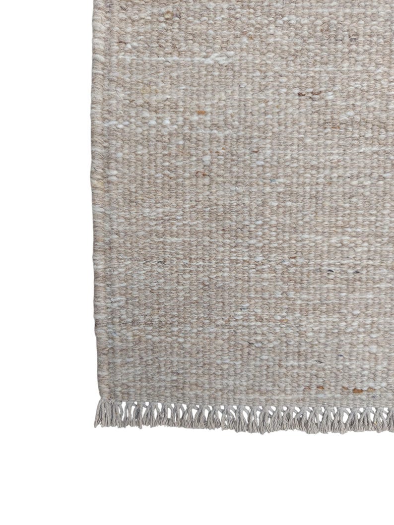 K&ouml;nnte beinhalten: Ein rechteckiger Webteppich in einer neutralen Farbpalette aus Beige und Off-White. Die Oberfl&auml;che zeigt ein gleichm&auml;&szlig;iges, dichtes Schlingenwebmuster mit dezenten Sprenkeln in Hellbraun und Creme, das mit einer kurzen Fransenborte abschlie&szlig;t.