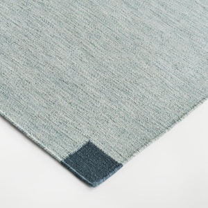 Könnte beinhalten: Ein rechteckiger Teppich mit einer strukturierten, hellblaugrauen Oberfläche und einem soliden, dunkelmarineblauen quadratischen Akzent an einer der Ecken. Das Gewebe wirkt dicht und gleichmäßig und erzeugt ein einheitliches Muster.