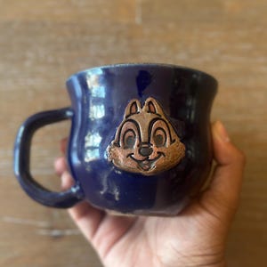 May include: A dark blue ceramic mug with a brown Chip character face on the front. The mug has a curved handle and a glossy finish. The mug is held in a person's hand.