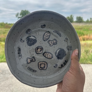 May include: A speckled grey ceramic bowl with a slightly irregular rim. The interior features black and tan food illustrations. The bowl is held by a hand against a backdrop of a field and cloudy sky.