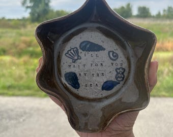 May include: A handmade, star-shaped ceramic dish with a brown exterior and a gray interior. The dish features blue seashell designs and the text "I'll wait for you by the sea."