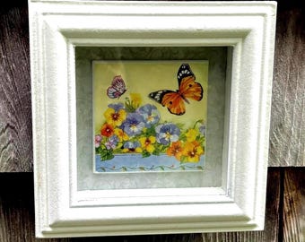 May include: A framed decorative tile featuring a floral and butterfly design. The artwork showcases colorful pansies and two butterflies against a yellow backdrop. The white frame adds a classic touch to the piece.