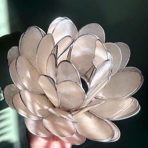 May include: A handcrafted, cream-colored flower sculpture with a layered petal design. The petals have a pearlescent sheen and are outlined with a silver-colored metal. The sculpture is a decorative art piece.