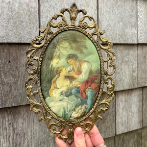 May include: An antique oval-shaped picture frame with a gold-colored, ornate metal frame. The framed artwork depicts a romantic scene with two figures in period clothing. The frame is held by a hand.