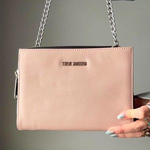 May include: A blush-colored clutch with a silver chain strap. The rectangular bag features a zipper closure and the brand name "STEVE MADDEN" in silver lettering. The bag is held by a person.