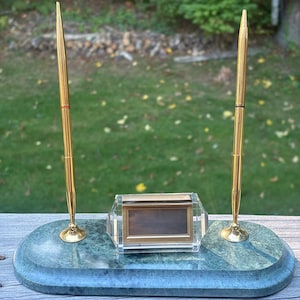 May include: A vintage desk set featuring a green marble base with two gold-colored pens and a small picture frame. The pens stand upright, and the frame is centered on the base. The set is designed for a desk.