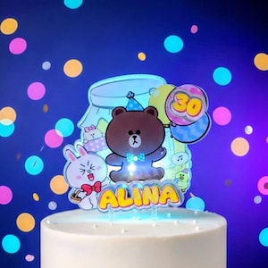 May include: A cake topper with a cartoon bear wearing a party hat and bow tie, holding its arms out. The topper has the name "ALINA" in yellow letters with a blue outline. Balloons with the number "30" are also included.