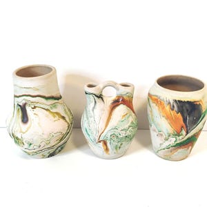 May include: Three ceramic vases with a marbled finish in shades of green, orange, black, and white. The two outer vases have wide openings, while the central vase features a double-spout design connected by a small arched handle at the top.