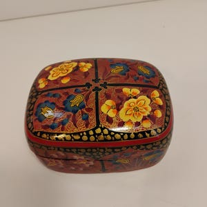 May include: A decorative, rectangular box with a hinged lid. The box is painted with a floral design in yellow, blue, and red on a dark red background. The edges are trimmed with black and gold accents.