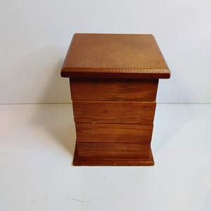 May include: A small, wooden box with a square top and four stacked, rectangular drawers. The box is made of a warm, medium-toned wood and sits on a white surface. The box is likely used for storage or display.