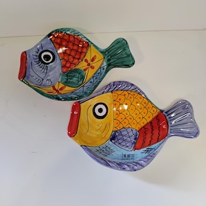 Puede incluir: Dos platos decorativos de cerámica con forma de pez. Los peces tienen un diseño colorido con detalles en rojo, amarillo, azul y verde. Cada pez tiene una boca roja, un ojo grande y una aleta caudal. Estos platos son probablemente para servir comida o como objetos decorativos.