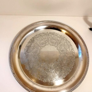 May include: A circular, silver-toned serving tray featuring an embossed floral pattern in the centre. The tray has a rope-style border. The tray is likely made of metal, and is suitable for serving food.