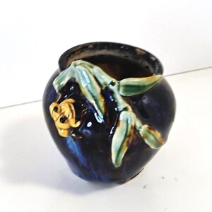 May include: A small, round, dark blue ceramic vase with a glossy finish. The vase is decorated with a relief design of bamboo leaves and a flower in shades of green and gold. The vase has a wide opening.