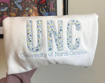 May include: A white sweatshirt with the letters "UNC" embroidered in a floral pattern of blue, yellow, and green. The words "University of North Carolina" are embroidered below the letters in a matching blue thread.