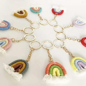 May include: A selection of rainbow keyrings. Each keyring has a woven rainbow design in various colours, including yellow, red, blue, and pink, with a white fringe. The rainbows are attached to gold-coloured key rings and chains.