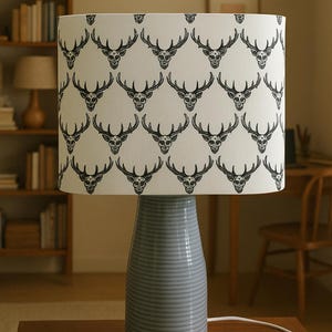 May include: A table lamp with a grey ceramic base and a white lampshade. The lampshade has a repeating pattern of black deer skull illustrations. The lamp is on a wooden table in a room with a bookshelf and other furniture. The base is approximately 30 cm tall.