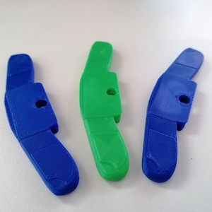 Könnte beinhalten: Drei 3D-gedruckte Kunststoffobjekte in Blau und Grün. Jedes hat eine gebogene Form mit einem rechteckigen Abschnitt und einem kreisförmigen Loch. Die Objekte sind auf einer weißen Oberfläche angeordnet.