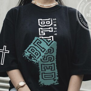 May include: Black t-shirt with the words "SIMPLY BLESSED" in white and teal block letters. A white cross is on the left sleeve. The shirt has a relaxed fit and short sleeves. Digital download available.