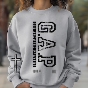 May include: Gray sweatshirt with a black graphic that says "GAP" and "GOD ANSWERS PRAYERS". The graphic is in a distressed font style. There is a small cross graphic below the text.