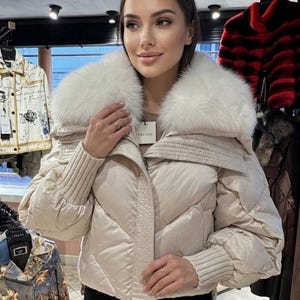 Könnte beinhalten: Beige Steppjacke mit großem weißem Pelzkragen. Die Jacke hat ein gestepptes Design und gerippte Bündchen. Das Etikett lautet "PRENSI". Die Jacke wird in einem Bekleidungsgeschäft ausgestellt.