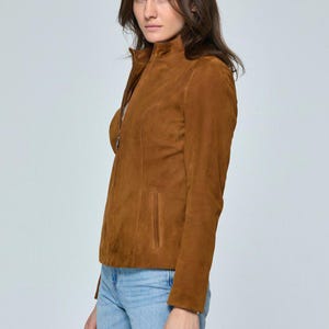 May include: A brown suede jacket with a high collar and a zipper closure. The jacket has a tailored fit and a small pocket on the front. The model is wearing light blue jeans.