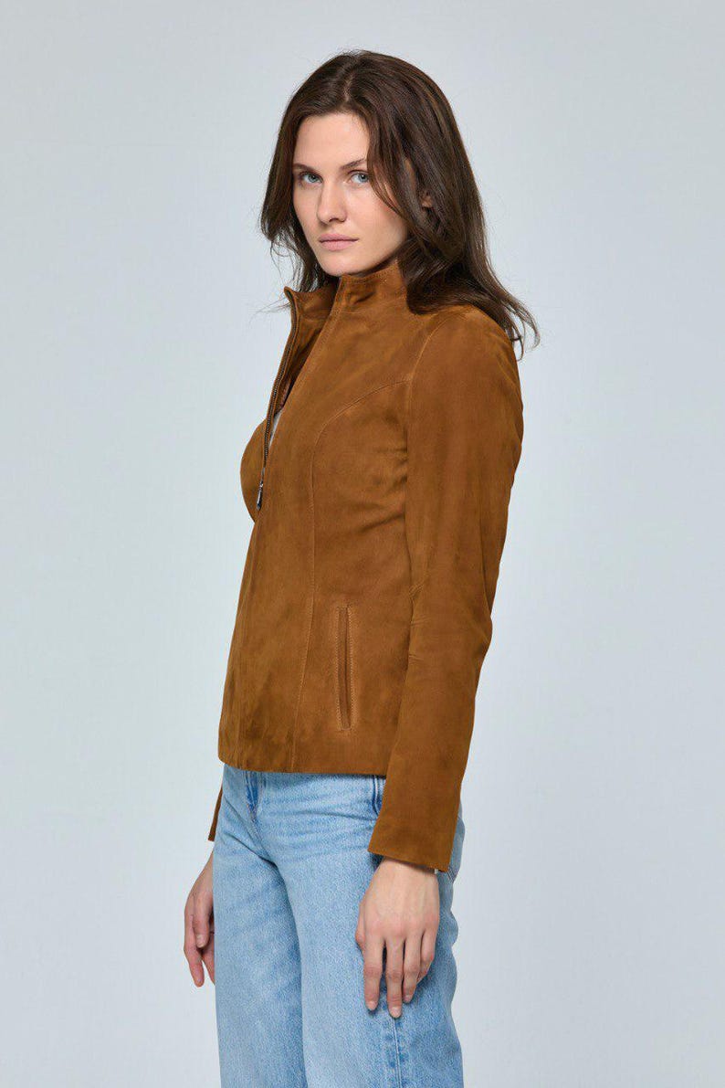 May include: A brown suede jacket with a high collar and a zipper closure. The jacket has a tailored fit and a small pocket on the front. The model is wearing light blue jeans.