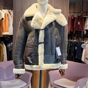 May include: A brown leather jacket with a cream-colored shearling lining and collar. The jacket features a front closure, two zippered pockets, and a large patch pocket. The jacket is displayed on a mannequin in a clothing store.